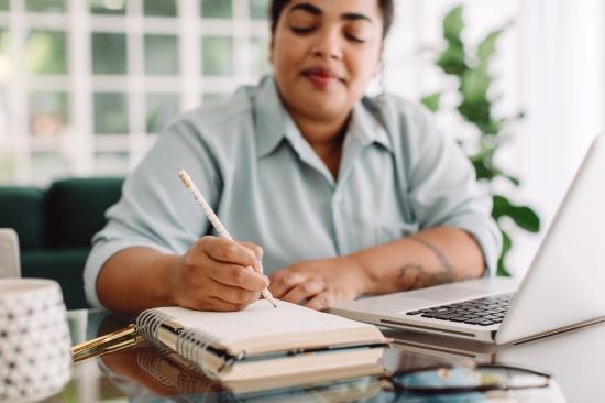 A person writing in a notebook beside a laptop