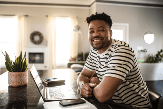 Smiling man sitting at a counter with a laptop in a bright home setting.